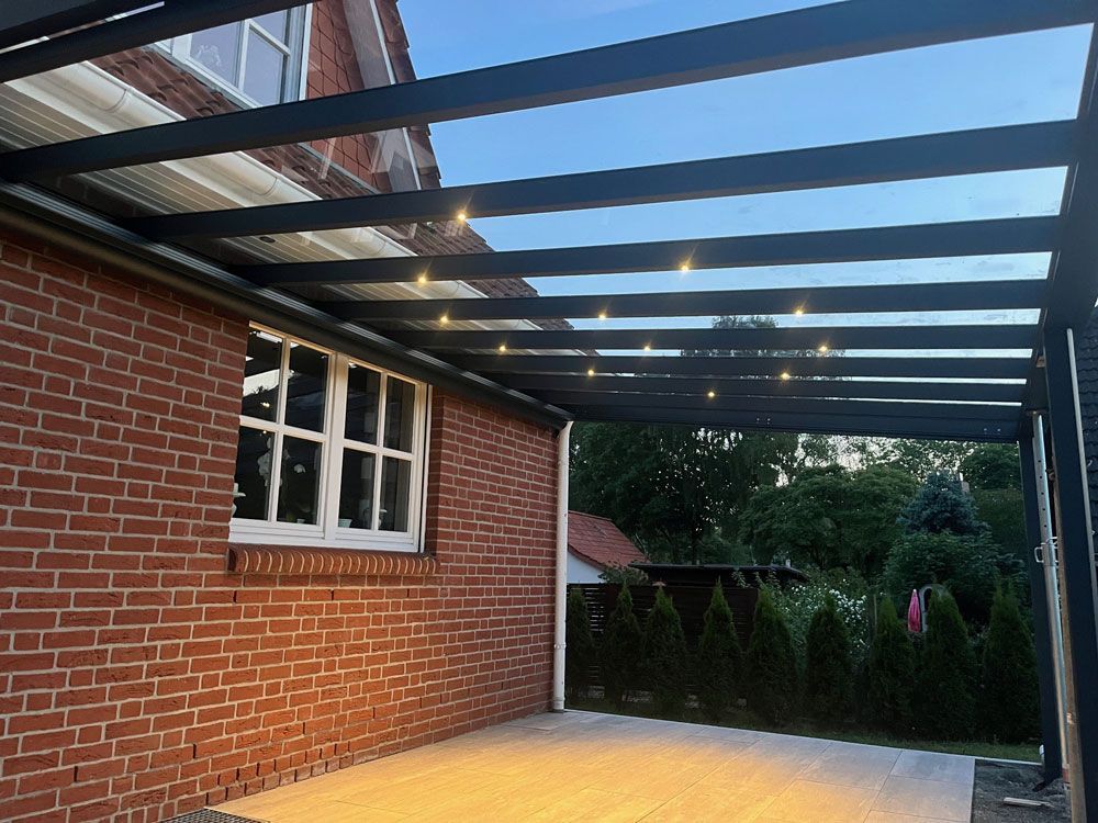 Terrasse mit transparentem Dach, Beleuchtung und Backsteinmauer. Abendhimmel sichtbar.