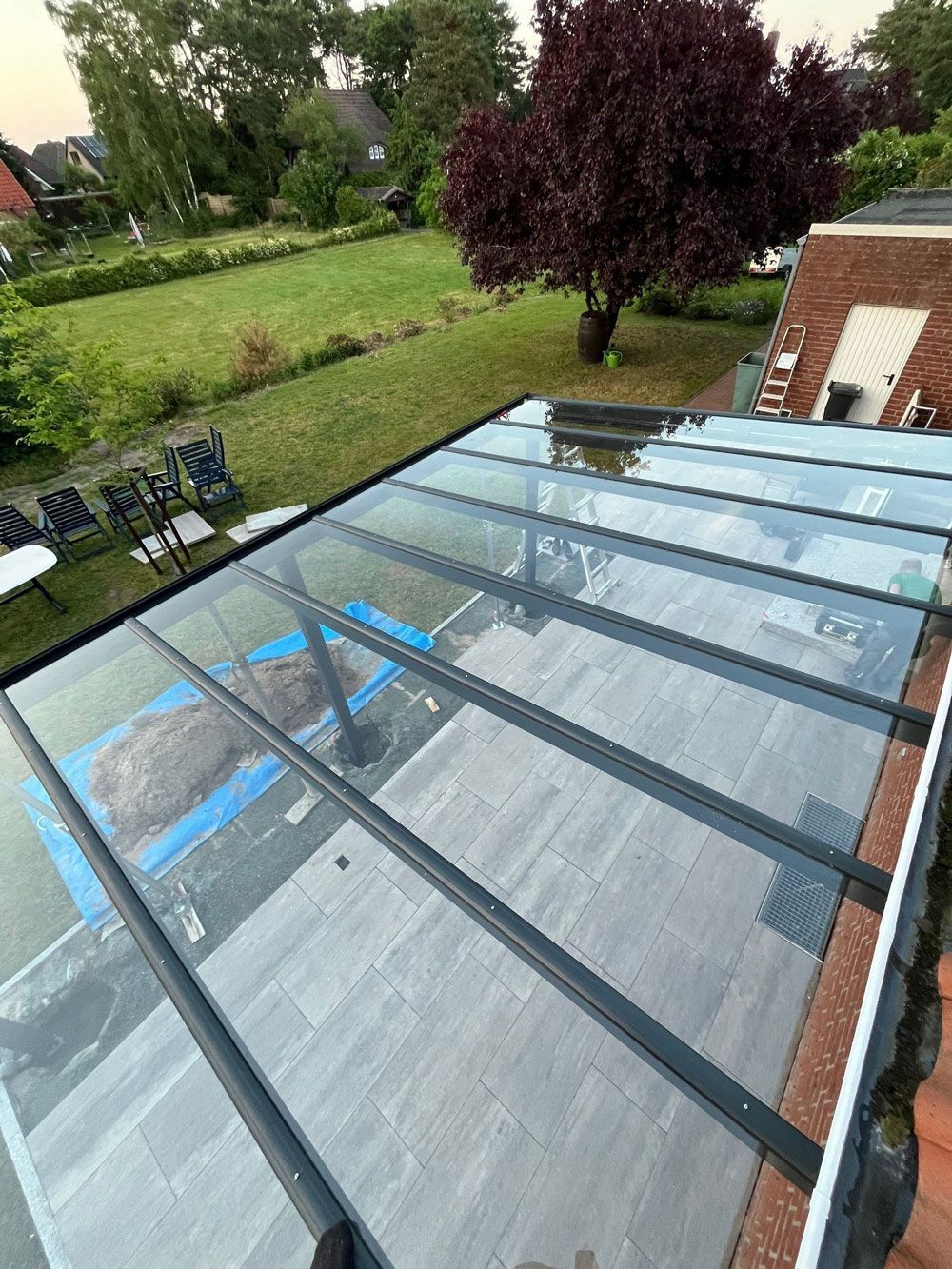 Terrasse mit Glasdach und Metallstützen, mit Blick auf einen Hinterhof mit grünem Rasen, Bäumen und Gartenmöbeln.