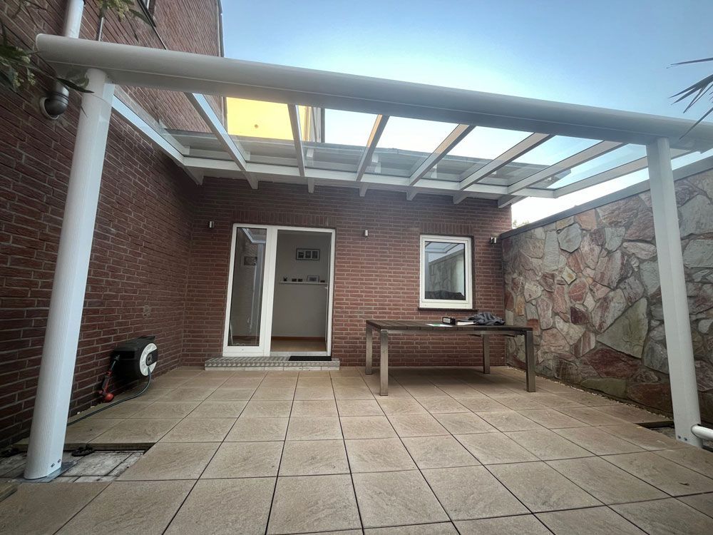 Terrasse mit Glasdach, Backsteinmauer, weißen Stützbalken und einem Holztisch.