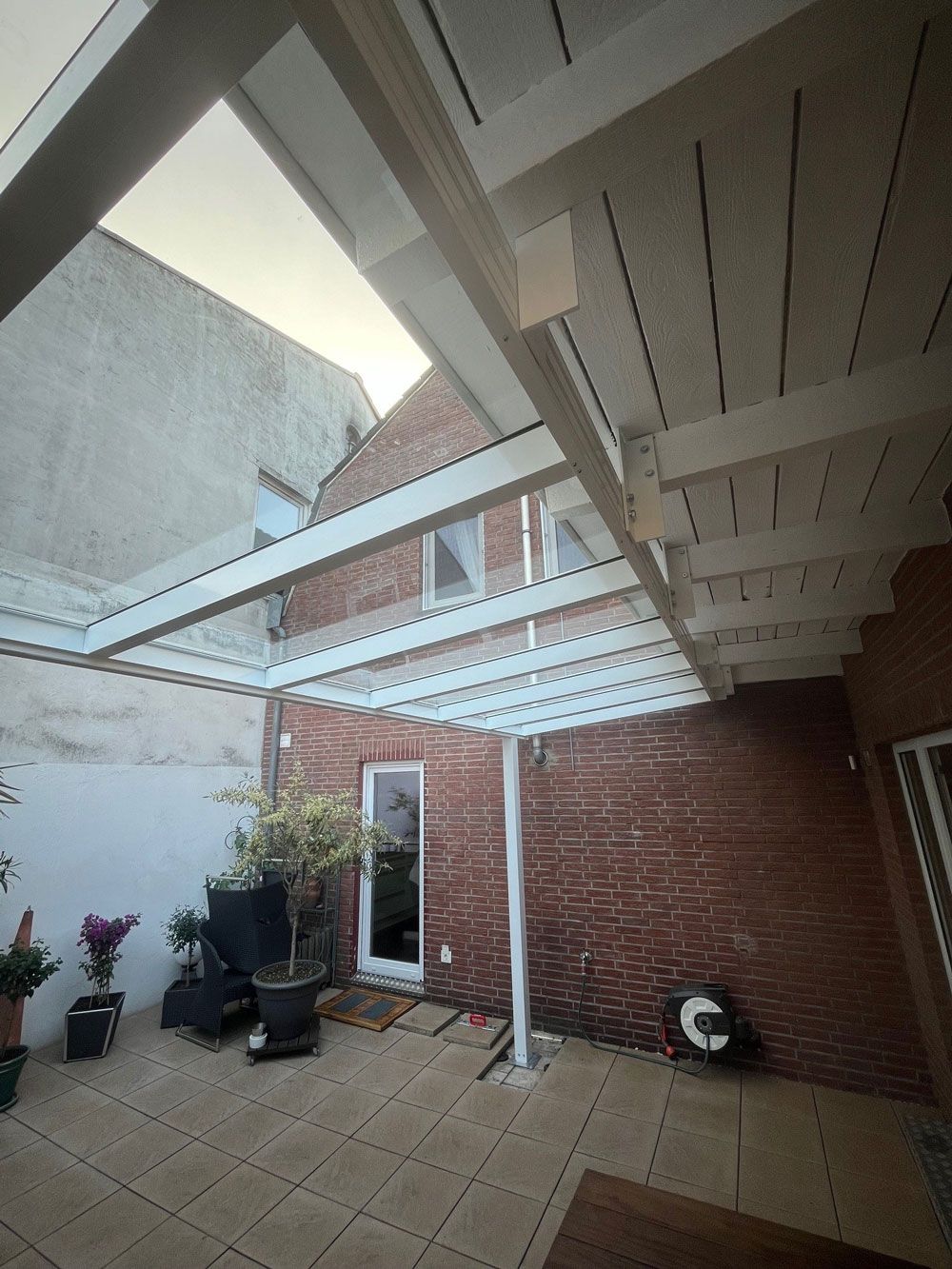 Terrasse mit weiß gerahmtem Glasdach und Backsteinmauer. Topfpflanzen auf der Terrasse.
