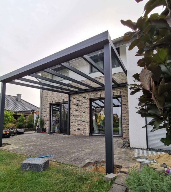 Schwarze Pergola mit Glasdach, die an ein Backsteinhaus über einer gepflasterten Terrasse angebaut ist.
