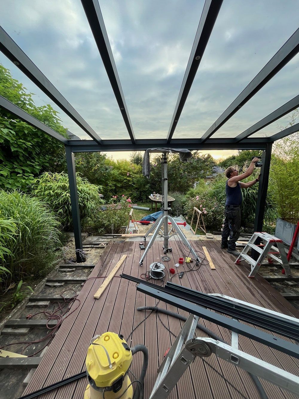 Ein Mann montiert eine schwarze Pergola mit Glasdach auf einer Holzterrasse in einem Garten.