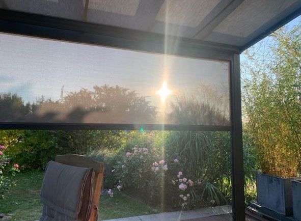 Sonnenlicht scheint durch einen teilweise heruntergelassenen Sonnenschutz im Freien; Grünpflanzen und ein Stuhl sind zu sehen.