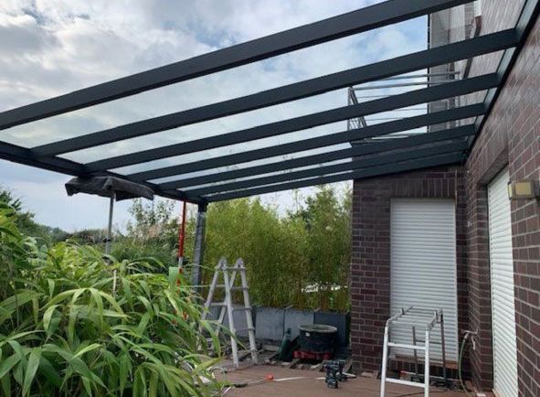 Terrasse mit Glasdach. Schwarzer Metallrahmen über transparenten Paneelen. Neben einem Backsteinhaus und Pflanzen.