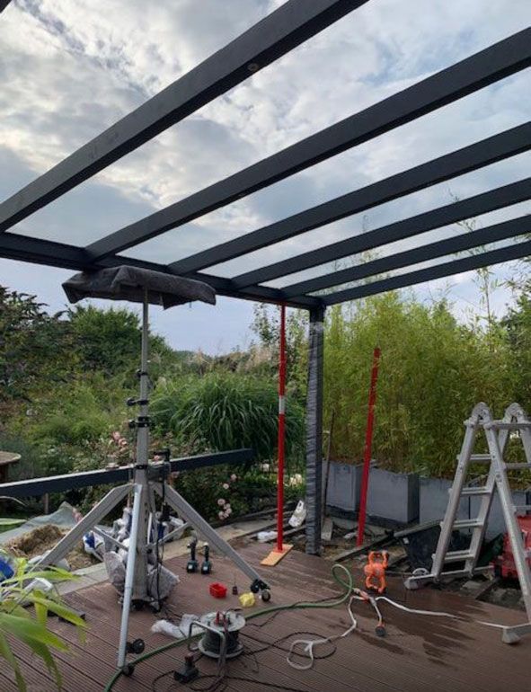 Eine Pergola wird auf einer Terrasse gebaut; im Hintergrund sind Werkzeuge, eine Leiter und Grünpflanzen zu sehen.