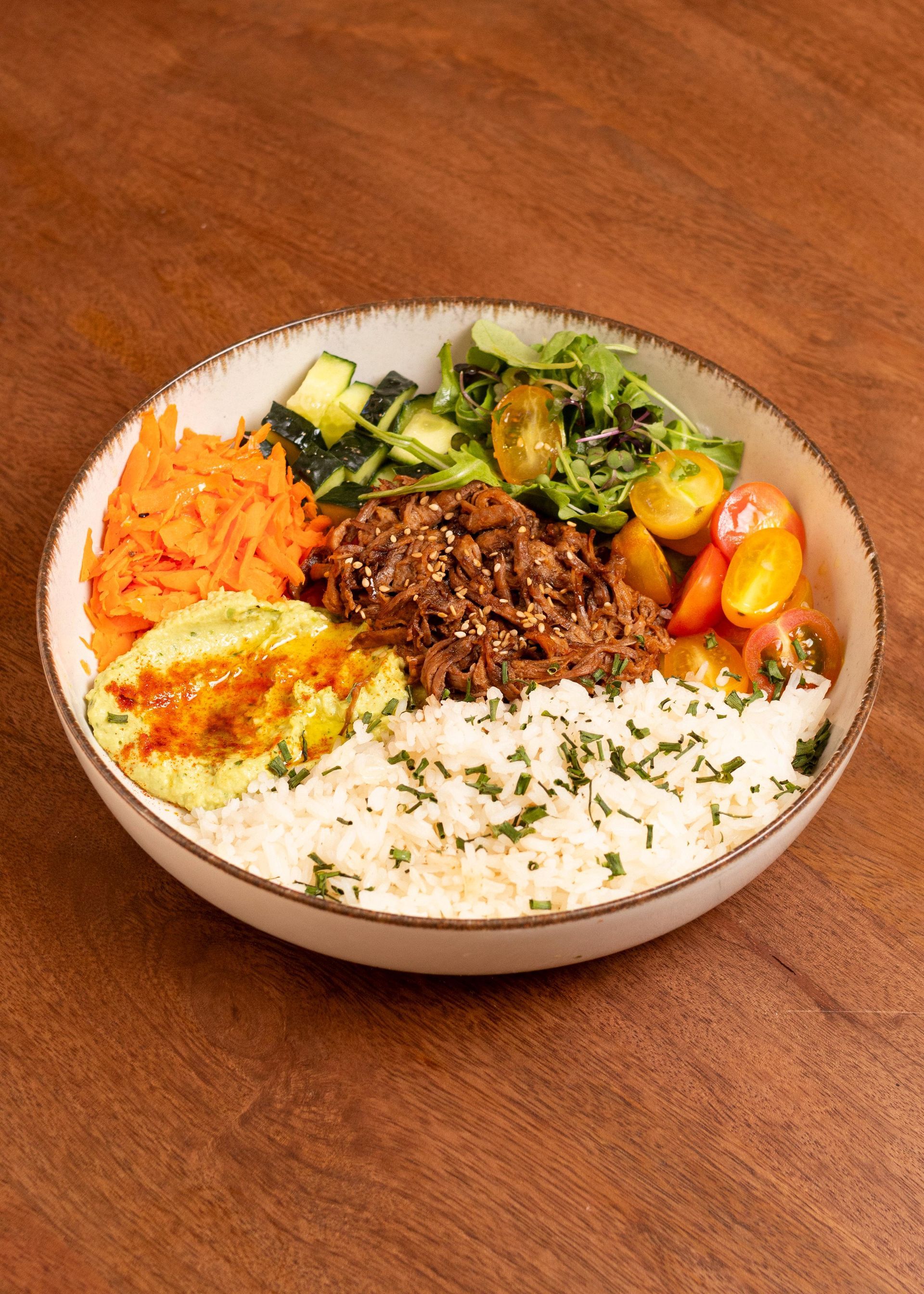 Un bol de arroz con carne picada, verduras y aguacate en rodajas sobre una mesa de madera.