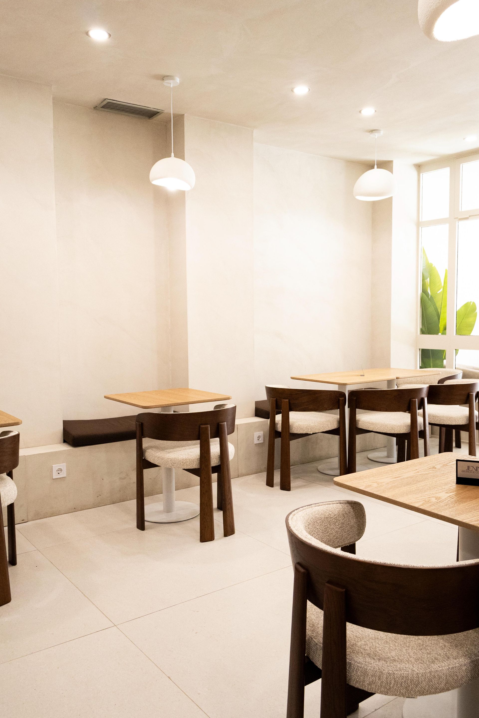 Interior luminoso de la cafetería con mesas y sillas de madera, lámparas colgantes y una ventana con plantas verdes.