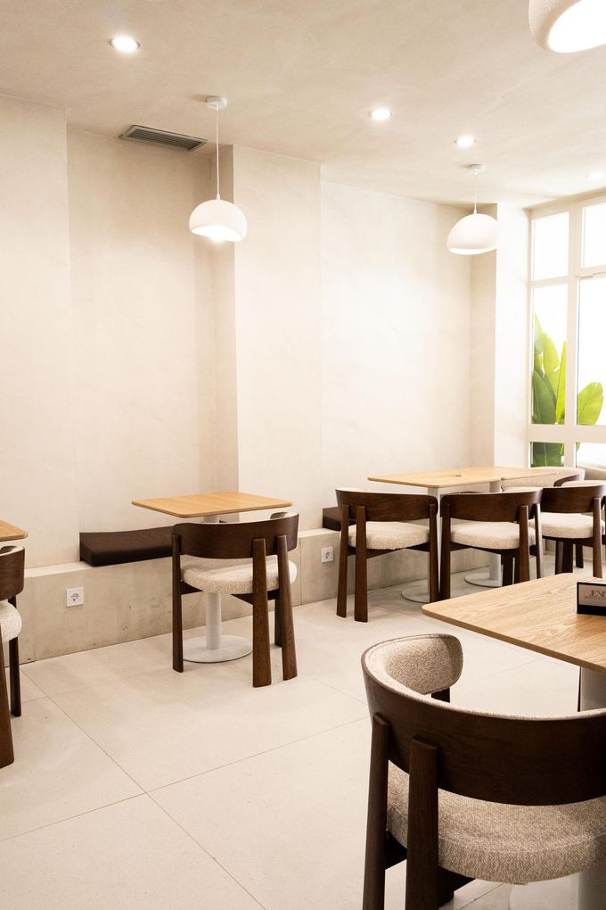 Interior luminoso de la cafetería con mesas y sillas de madera, lámparas colgantes y una ventana con plantas verdes.