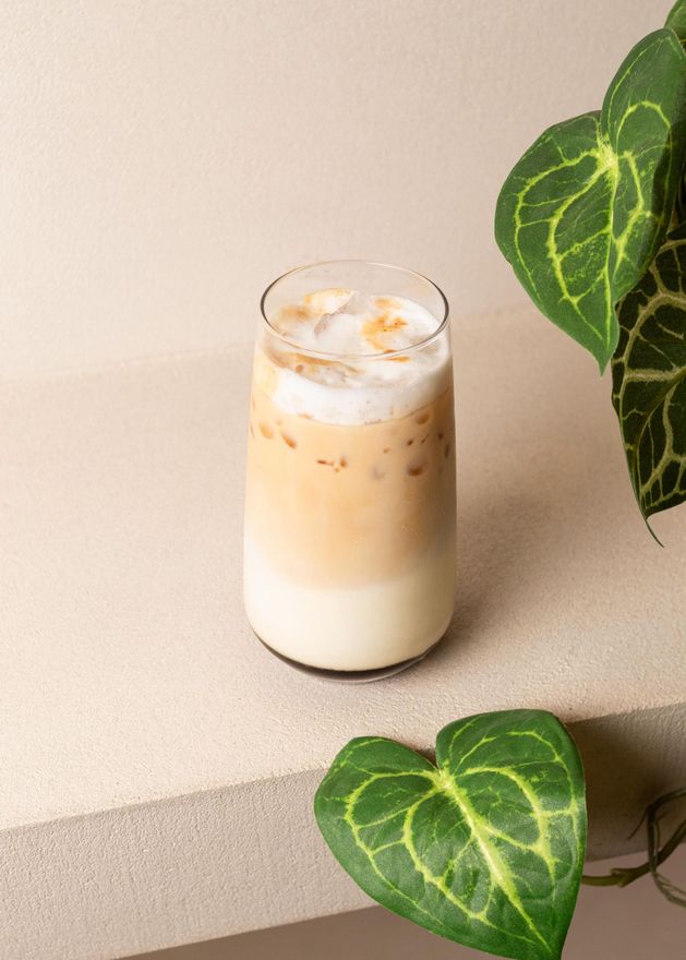 Un café con leche helado en un vaso sobre una repisa beige, enmarcado por hojas verdes en forma de corazón.
