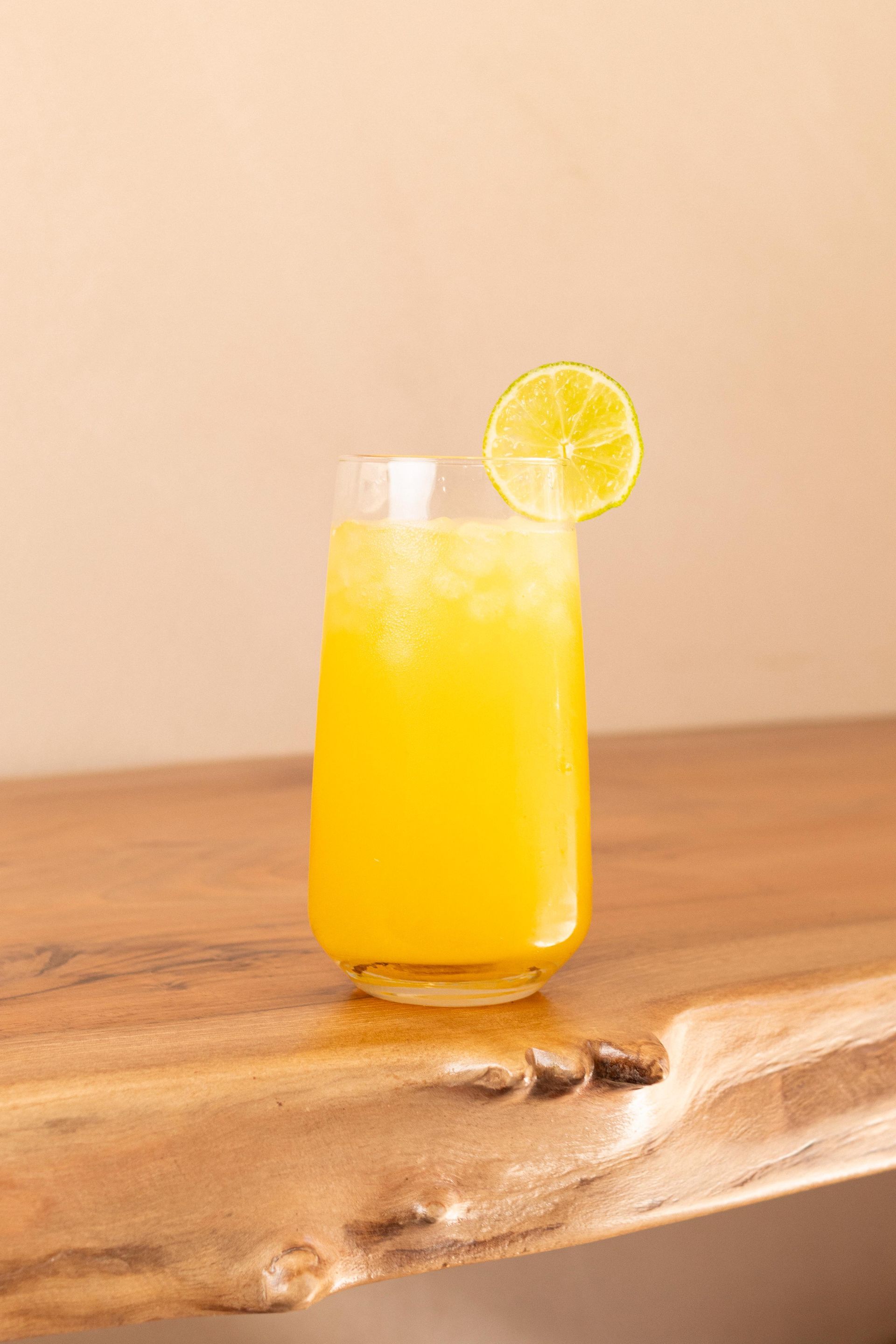 Una bebida cítrica amarilla en un vaso alto con una rodaja de limón, sobre una mesa de madera.
