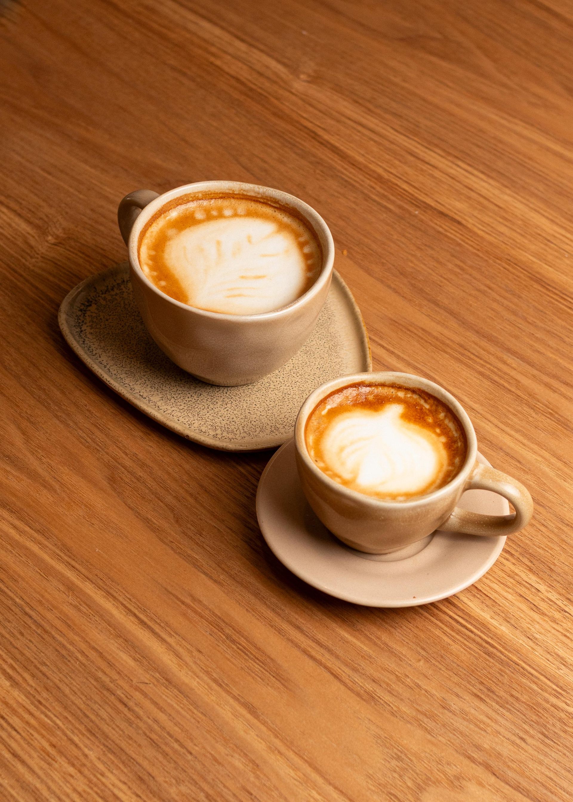 Dos tazas de café con leche sobre sus platillos encima de una mesa de madera, vistas desde arriba.