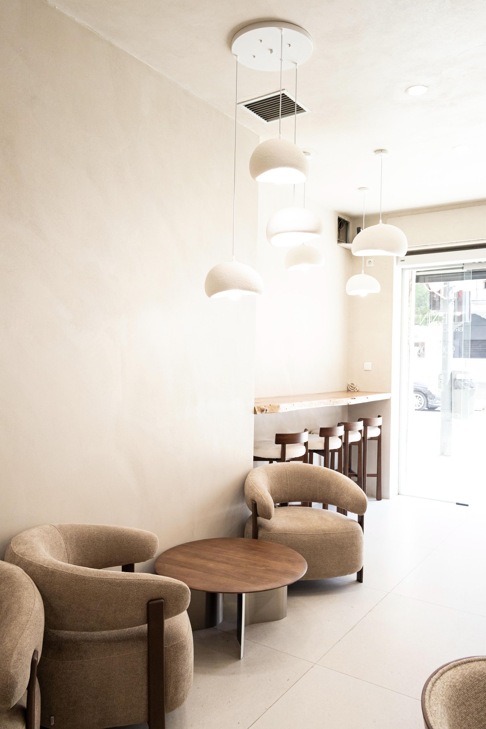 Cafetería minimalista con mesas redondas, sillas de mimbre, lámparas colgantes y una entrada luminosa.