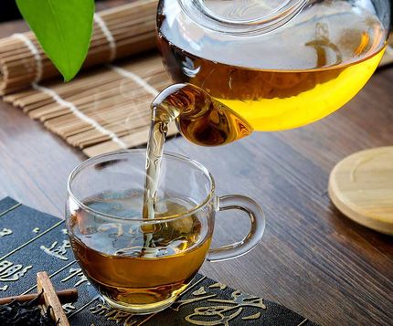 Té ámbar vertiéndose de una tetera de cristal a una taza transparente sobre una mesa de madera.
