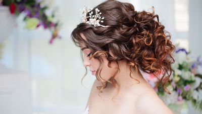 Frau mit lockigem braunem Haar, Hochsteckfrisur und Krone, Blick nach unten, Blumen im Hintergrund.