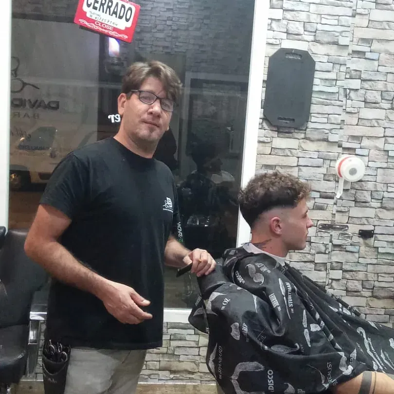 Un hombre con una camisa negra está de pie junto a un hombre que se corta el pelo frente a un cartel que dice cerrado