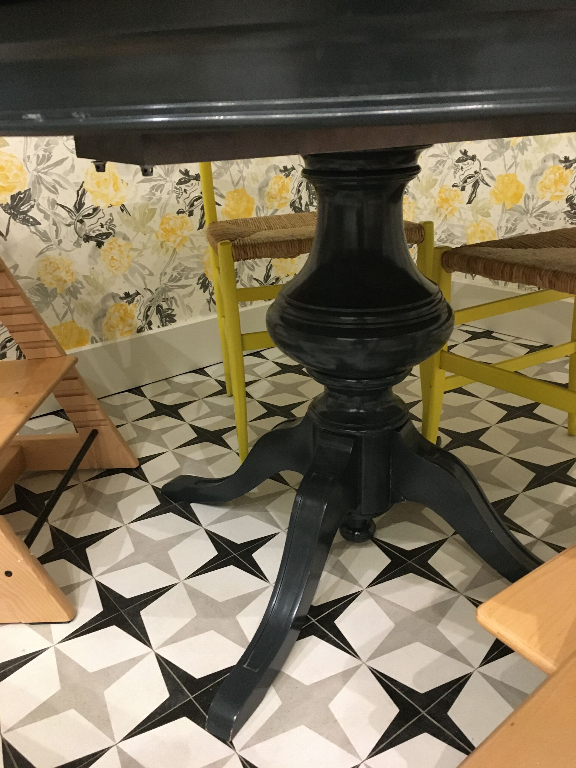 Mesa de pedestal azul oscuro sobre suelo estampado, sillas amarillas y papel tapiz floral de fondo.