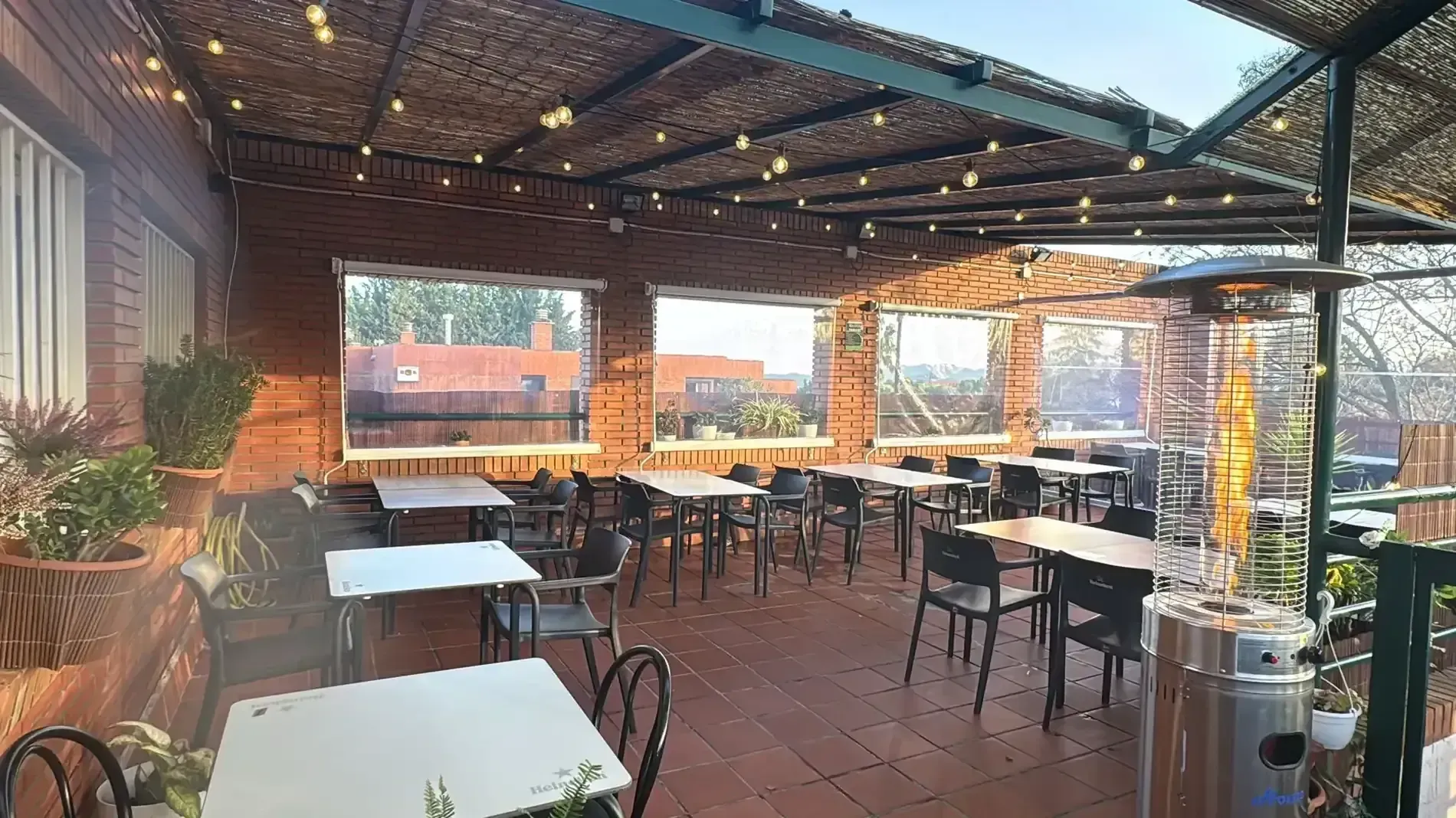 Patio de restaurante al aire libre con mesas, sillas y guirnaldas de luces. Estructura de madera y ventanas.