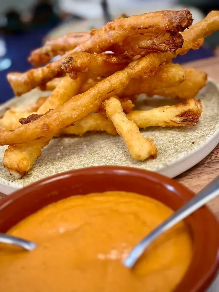 Palitos de verduras fritas apilados en un plato, servidos con un tazón de salsa de naranja.