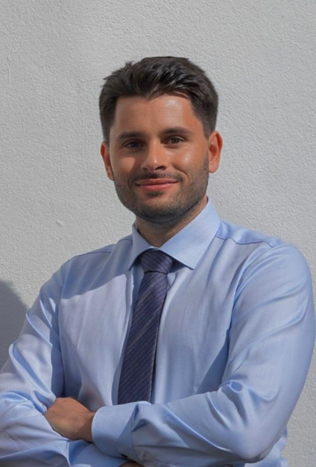 Eine Person in einem hellblauen Button-Down-Hemd und einer lila gemusterten Krawatte steht mit verschränkten Armen an einer weißen Wand.