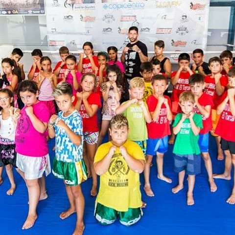 Grupo de niños en un gimnasio, posando en posición de boxeo. Colchoneta azul, con un ring de boxeo y pancartas al fondo.