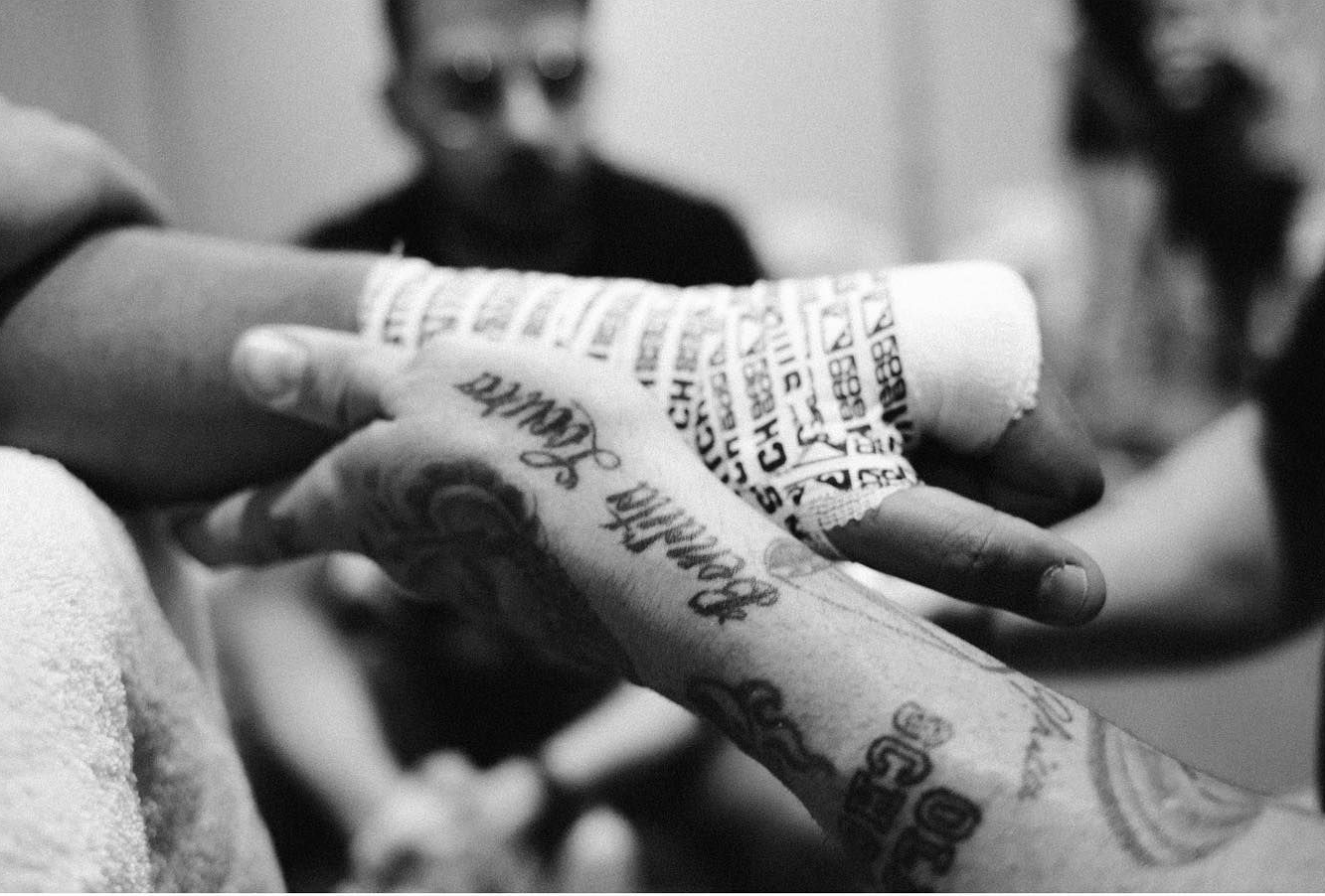 Una mano vendada con cinta deportiva blanca, preparándose para una pelea. Otra persona al fondo.