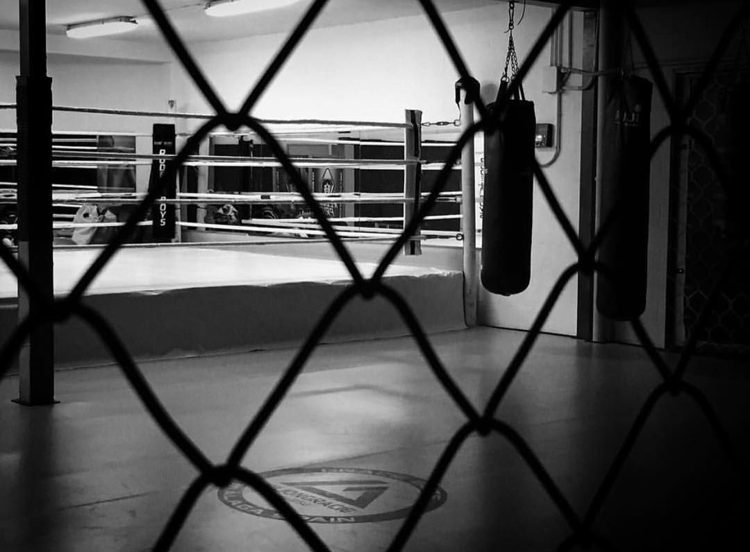 Ring de boxeo visto a través de una valla metálica, con sacos de boxeo colgados. Blanco y negro.