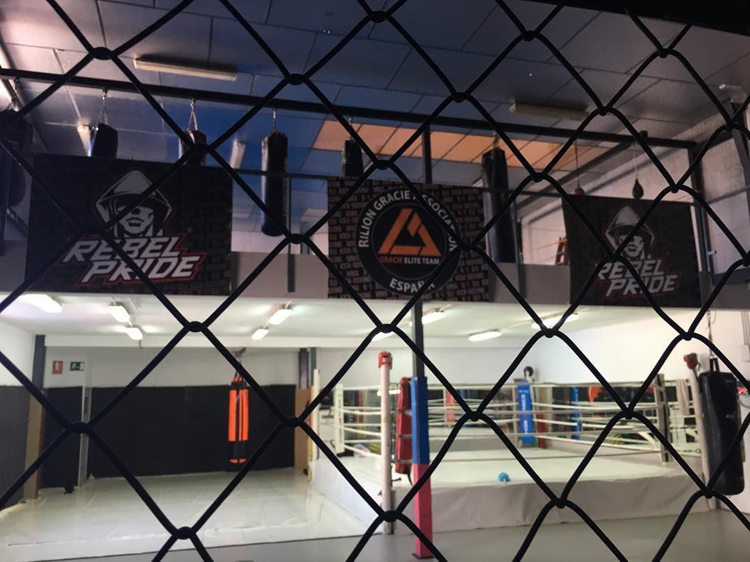 Interior de un gimnasio de boxeo, con un ring en el centro, sacos de boxeo y pancartas detrás de una valla de alambre.