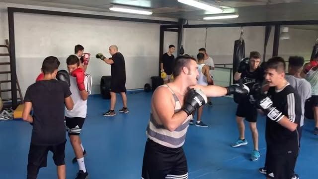 Clase de boxeo en un gimnasio; varias personas entrenan, una de ellas lanza un puñetazo. Suelo azul, guantes negros.