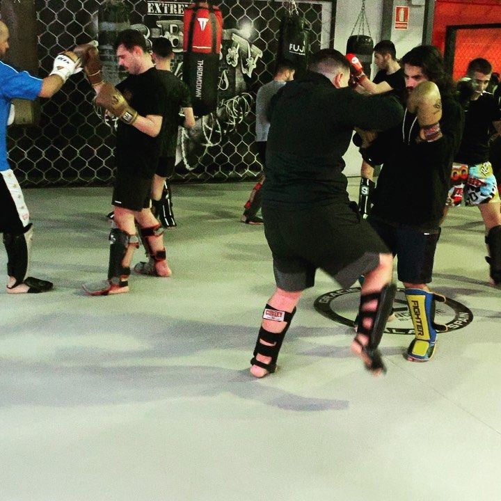 Personas entrenando en un gimnasio de artes marciales usando equipo de protección, pateando y golpeando.