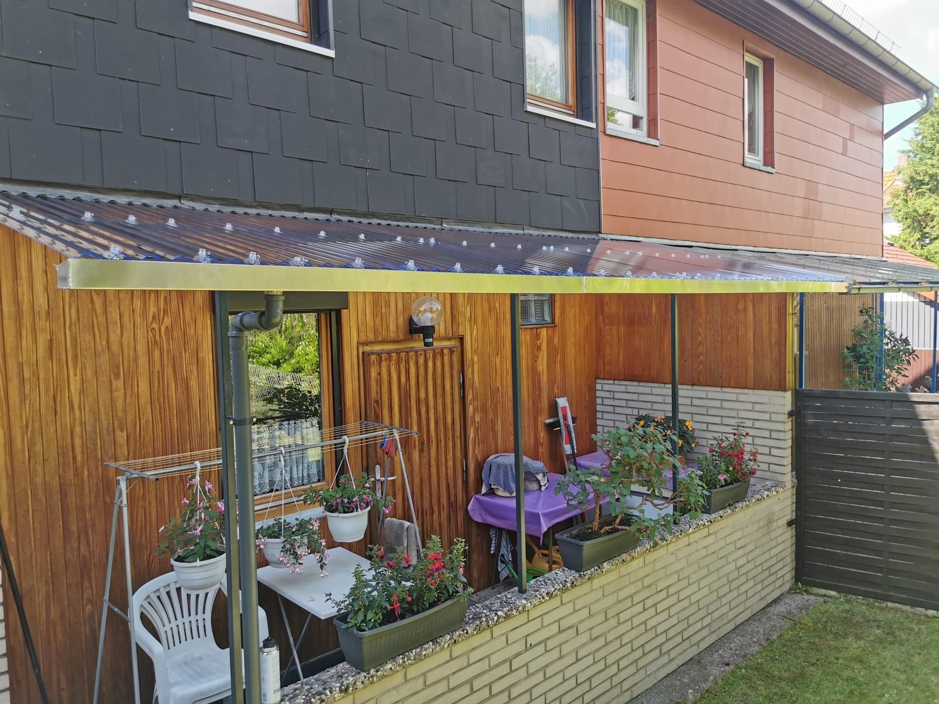 Eine Terrasse mit Tisch und Stühlen unter einem Vordach vor einem Haus.