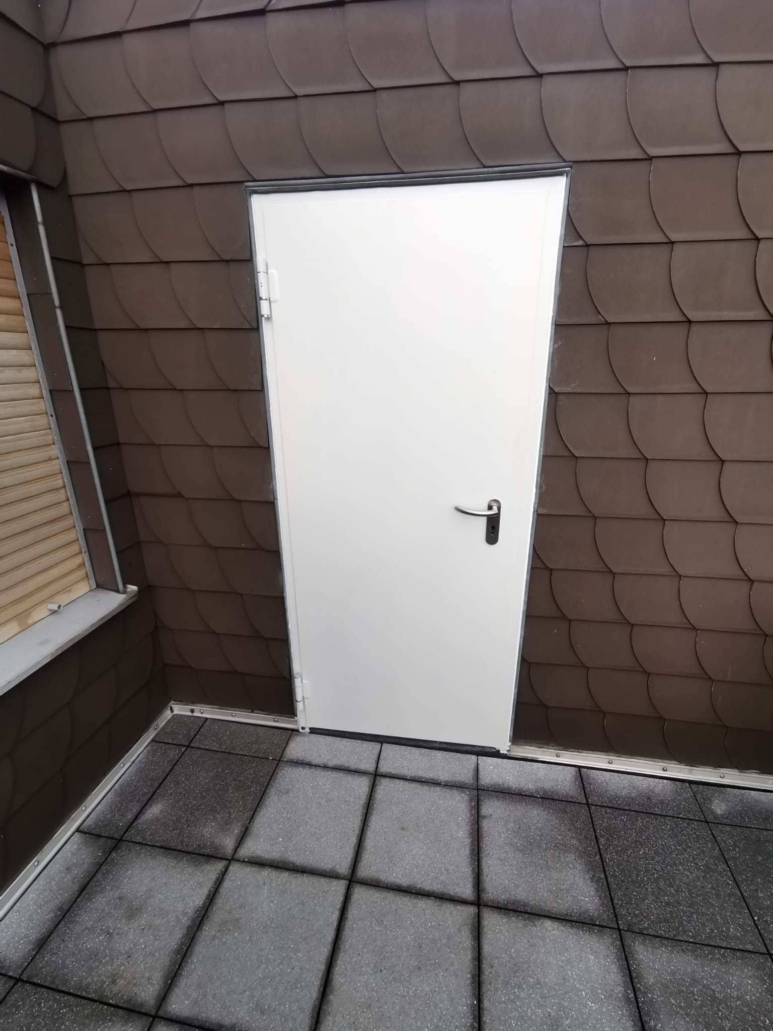 Auf einem gefliesten Boden steht neben einem Fenster eine weiße Tür.