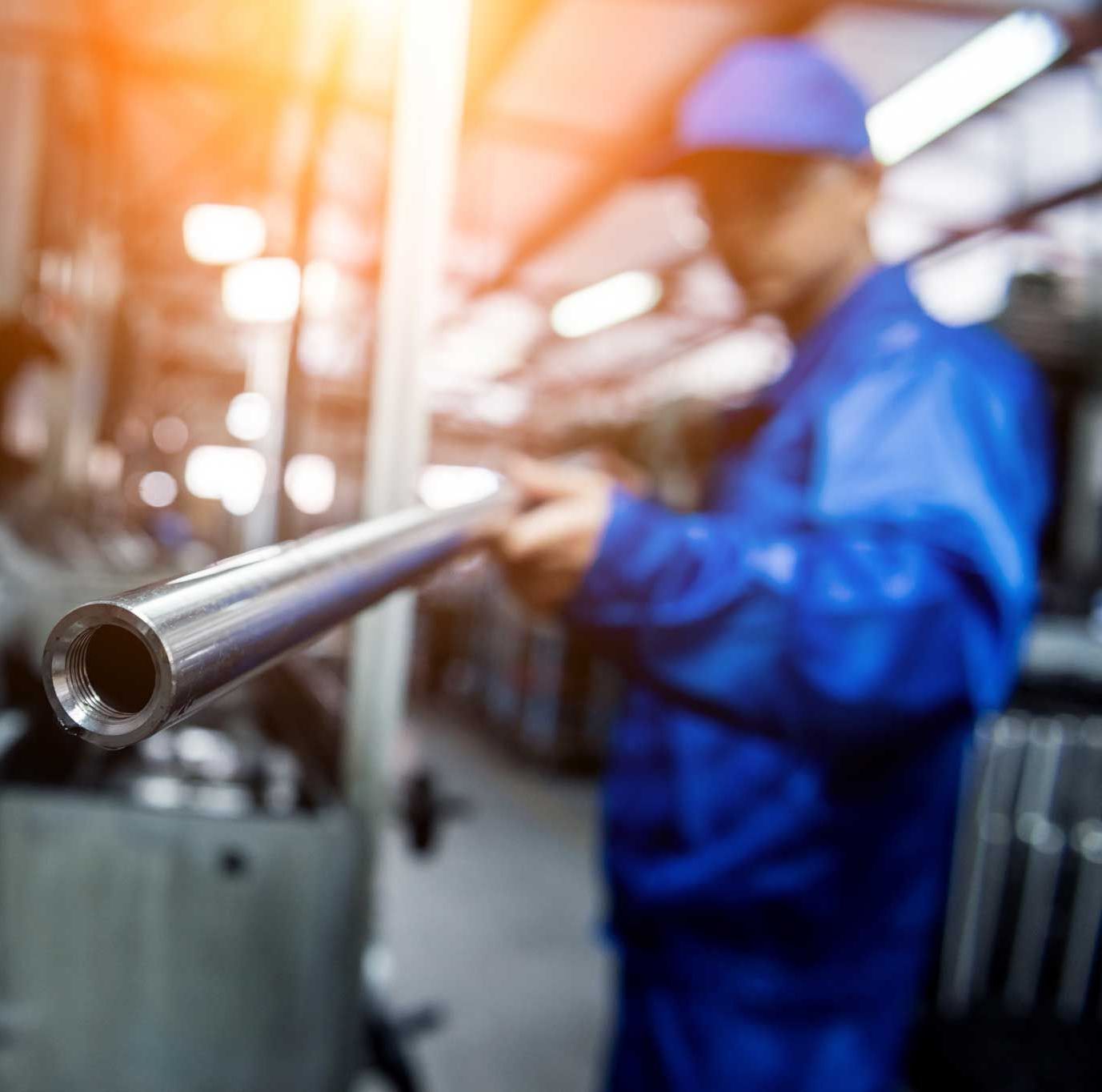 Ein Mann in einem blauen Overall hält in einer Fabrik ein Metallrohr.