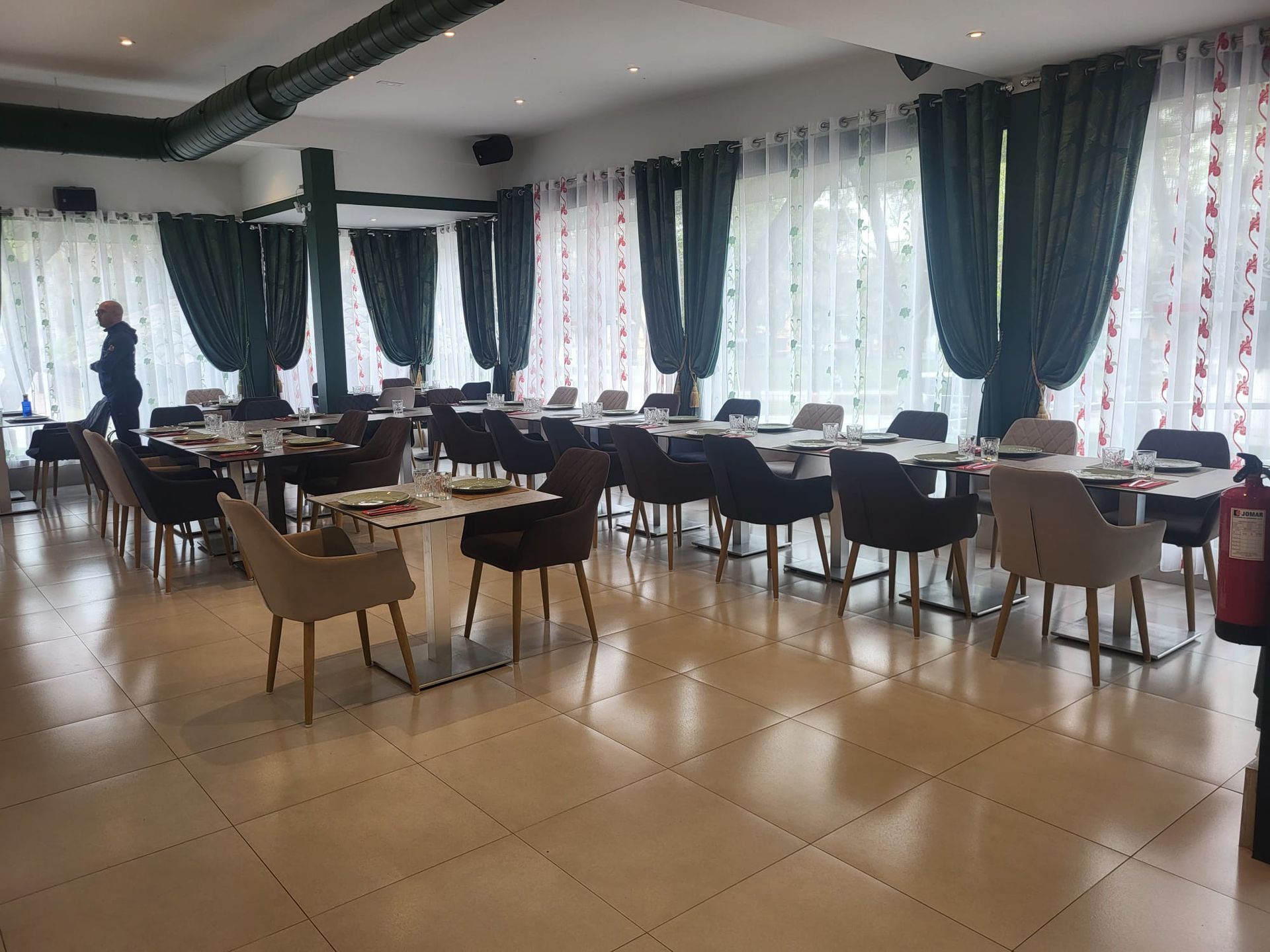 Interior de restaurante con mesas, sillas, cortinas verdes y una persona caminando al fondo.