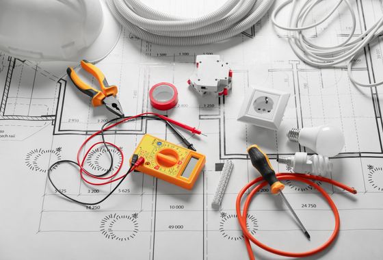 Des outils électriques, des fils électriques, un multimètre et une ampoule disposés sur des plans de construction.