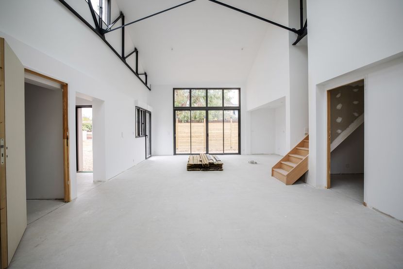 Vue intérieure d'une pièce lumineuse aux murs blancs, en construction, avec un haut plafond, des poutres apparentes et un escalier en bois.