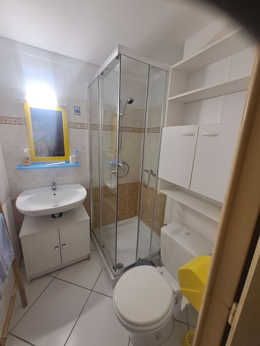 Petite salle de bains carrelée de blanc, comprenant une cabine de douche en verre, un lavabo sur pied avec un miroir à cadre jaune et des toilettes.