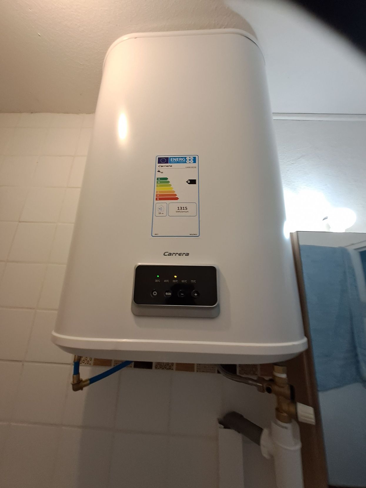 Un chauffe-eau électrique vertical blanc, fixé sur un mur carrelé, doté d'un panneau de commande numérique et d'une étiquette énergétique.