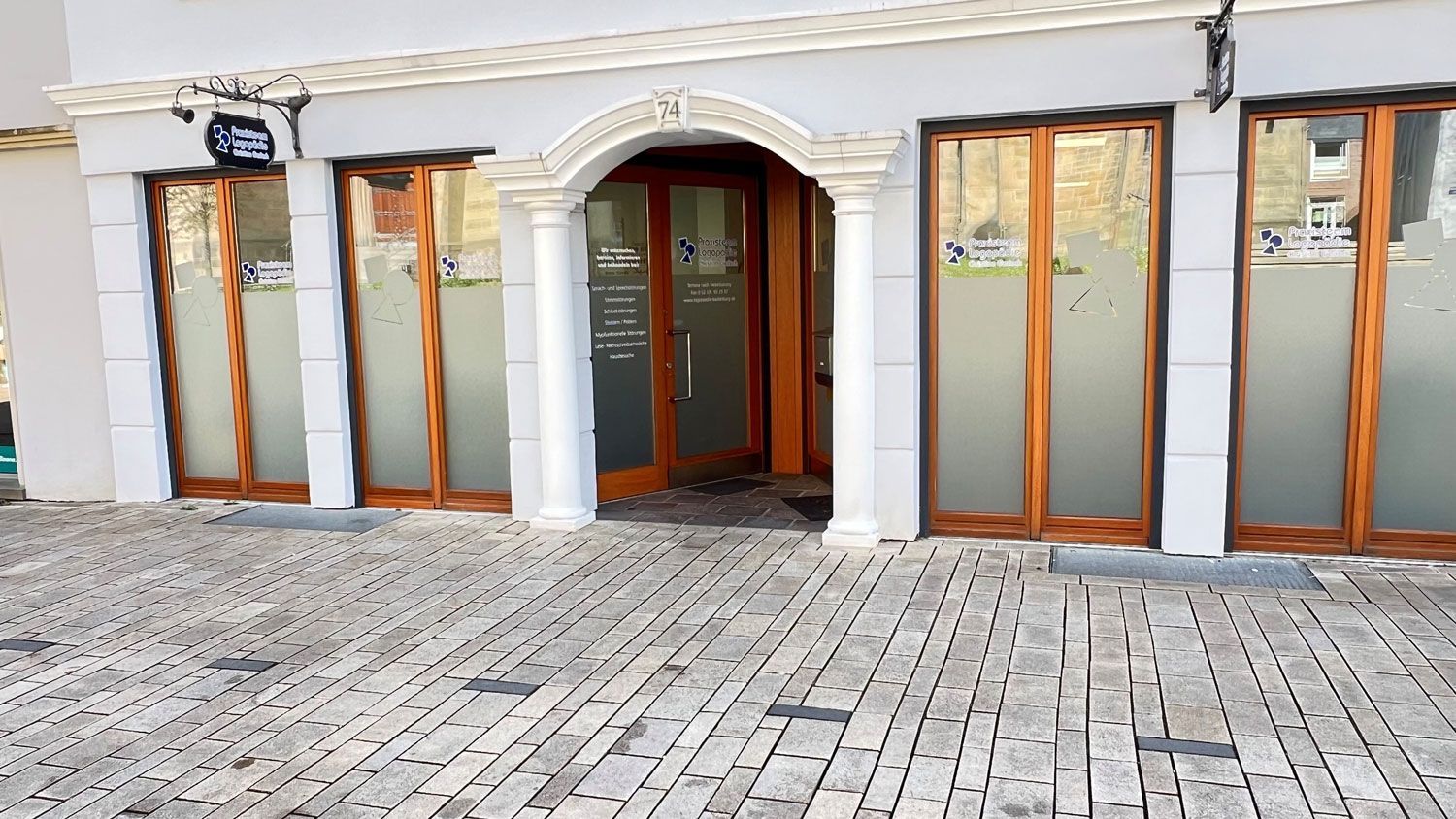 Außenansicht eines Gebäudes mit holzgerahmten Glastüren und einem zentralen, gewölbten Eingang. Kopfsteinpflaster.