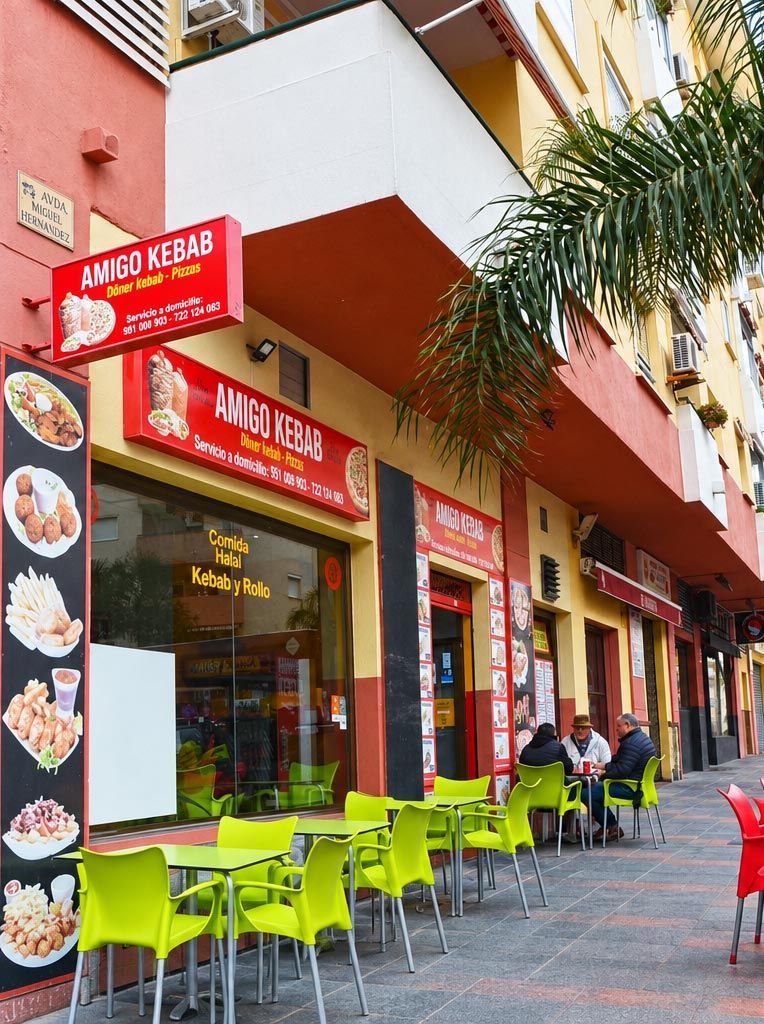 Colorido restaurante al aire libre con toldos rojos, sillas verde lima y mesas en la acera.