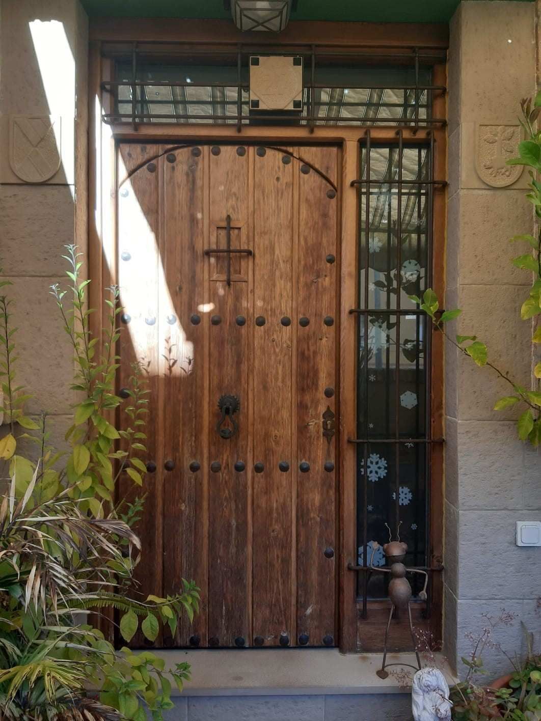 Una puerta de madera arqueada con remaches de hierro, una cruz y una reja vertical en la ventana, todo ello integrado en una fachada rústica de piedra.