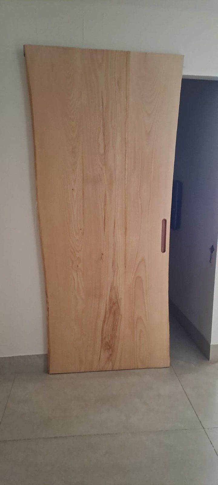 Una puerta corredera de madera clara con tirador vertical instalada contra una pared blanca y un suelo de baldosas de color claro.