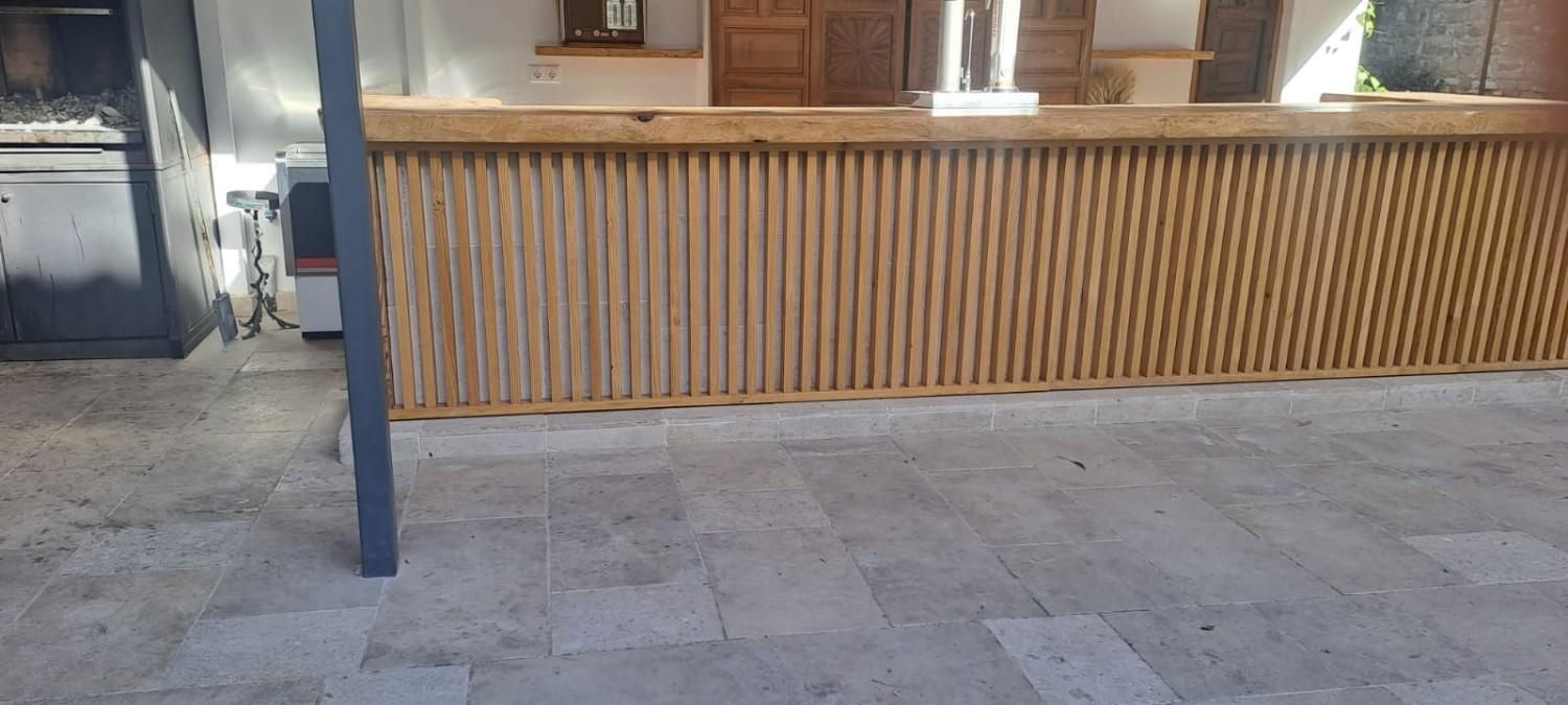 Una barra de madera con listones verticales instalada sobre un patio de piedra de color claro, situada junto a una parrilla exterior.