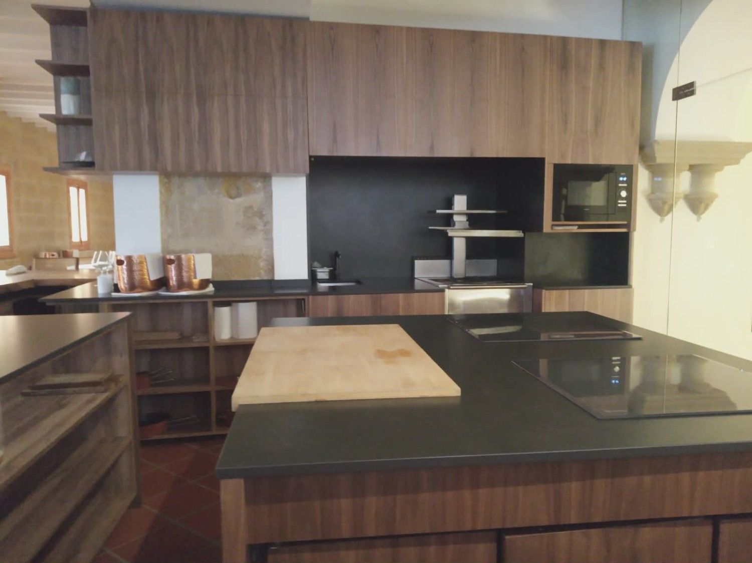 Una cocina moderna y minimalista con armarios de madera oscura, una gran isla con placa de cocción negra y microondas integrado.