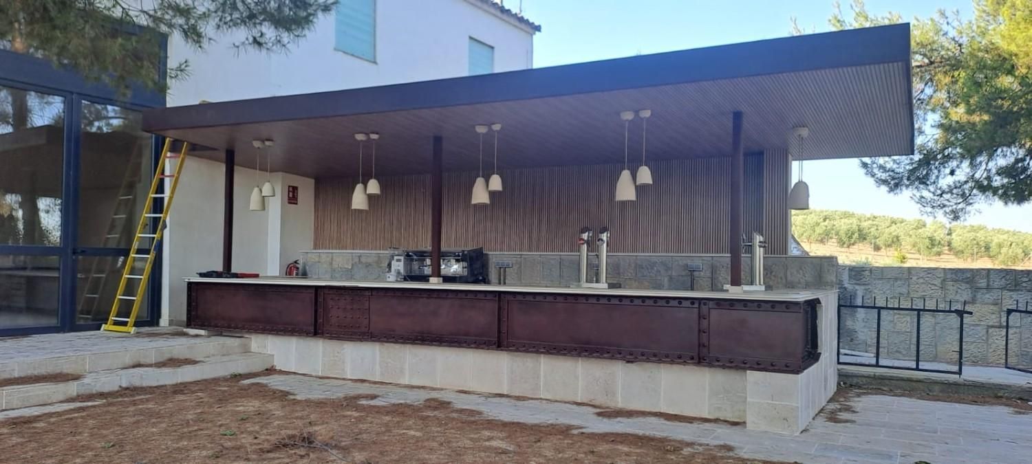 Bar al aire libre con barra de metal oscuro, base de piedra clara y techo de madera, con lámparas colgantes y una escalera.