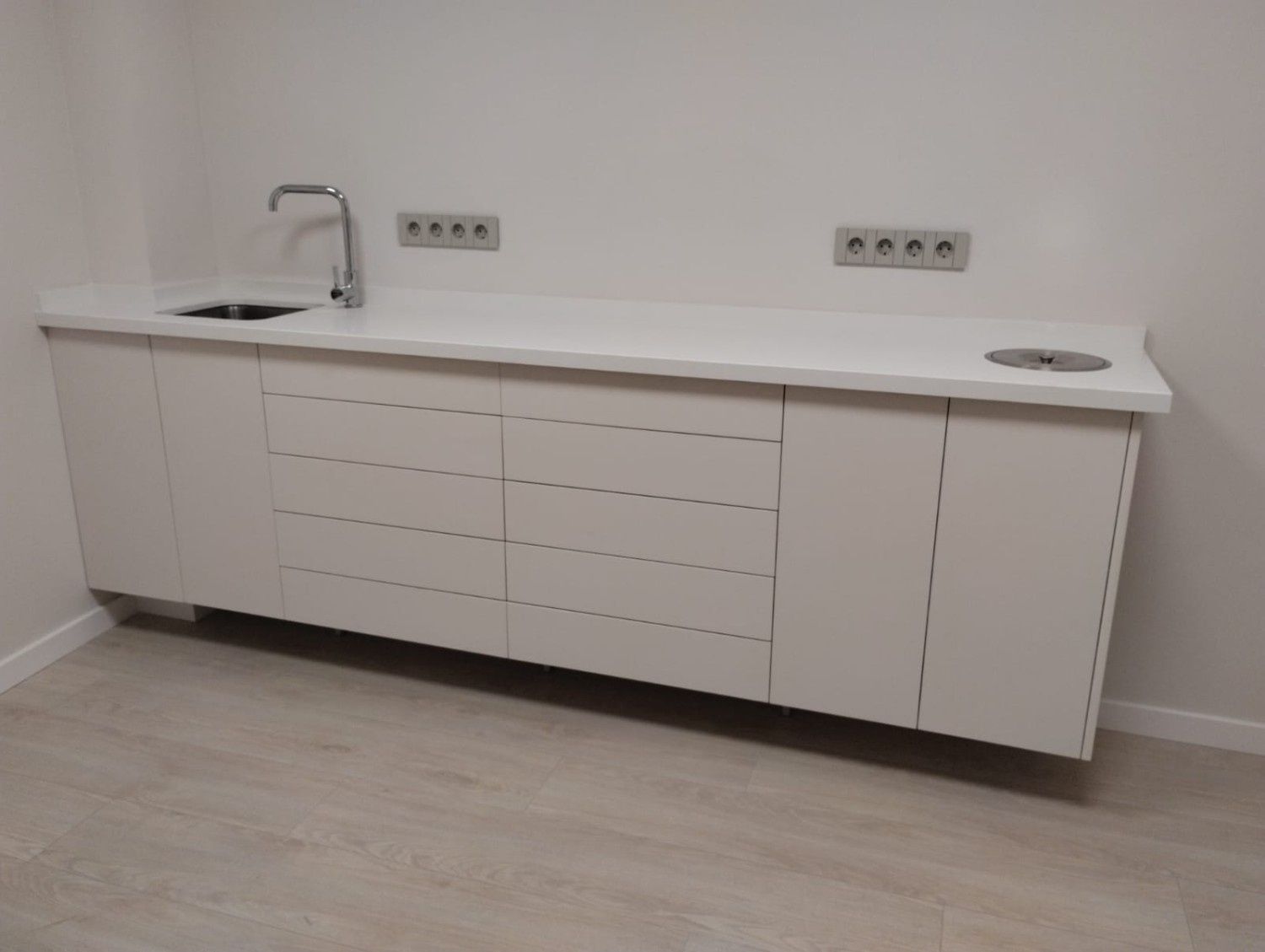 Un armario de servicio blanco, montado en la pared, con fregadero, grifo, cajones y conducto de residuos en una habitación minimalista.