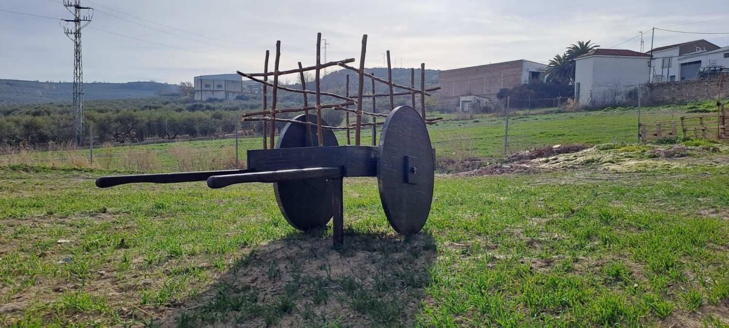 Un carro rústico de madera con ruedas grandes y sólidas descansa sobre un campo de hierba bajo un cielo brillante, con casas a lo lejos al fondo.