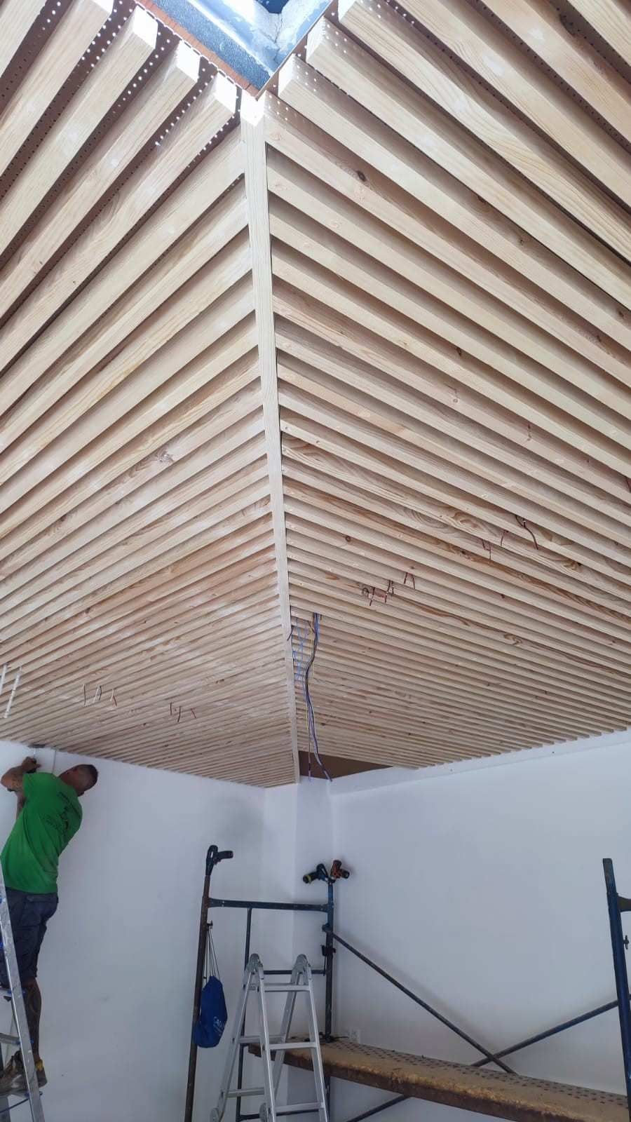 Un obrero subido a un andamio instala un techo de listones de madera en una habitación de paredes blancas.