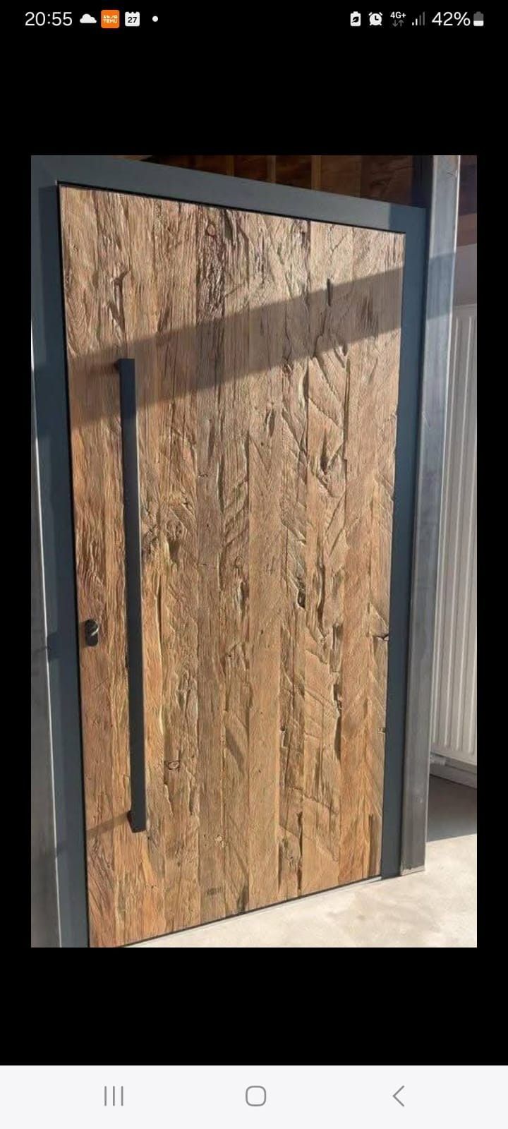 Una puerta de entrada moderna con manija vertical de metal oscuro y un panel de madera texturizada de color marrón claro con marco de metal.