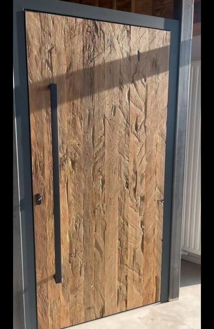 Una puerta de madera clara con textura, marco de metal oscuro y tirador largo y vertical de color negro.