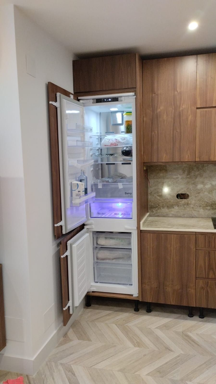 Un frigorífico alto empotrado con puertas que se abren, ubicado en una cocina moderna con armarios con acabado de madera y suelos de espiga.